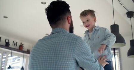 Low angle happy cute little son enjoying fun jumping into young fathers arms from house kitchen bar table slow motion.の写真素材