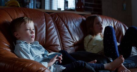 Entertainment and addiction, cute little Caucasian boy and girl watch TV at home on the couch with snacks slow motion.の写真素材