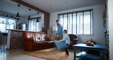 Happy young Caucasian father and little son playing, fighting pillows in modern light house living...