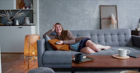 TV addiction on quarantine. Young happy attractive Caucasian blonde woman watching serial at home on comfortable sofa.の写真素材