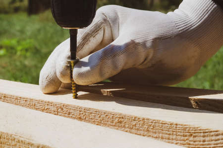 A hand with a screwdriver twists a screw into a wooden board. Construction