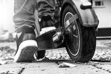 An electric scooter stands on the road strewn with autumn foliage. Eco transport. The leg of a young man stands on an electric scooter. black and white photoの写真素材