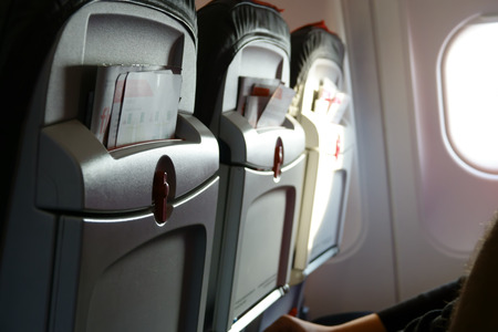 Three airplane seats with promotional brochures in aircraft passenger cabin. Light from porthole....