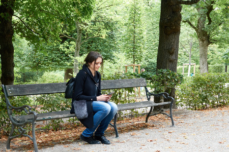 A girl in the park sits alone on a bench with a phone.の写真素材