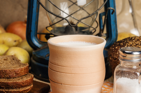 Milk in ceramic ware, rye bread, salt shaker with salt, vegetables and kerosene lantern on a wooden table. Still-life in the rustic styleの写真素材
