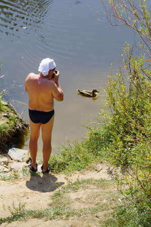 Arkaim, Chelyabinsk region, Russia - August 24, 2021: A man in swimming trunks photographs a wild duck standing on the riverbankのeditorial素材