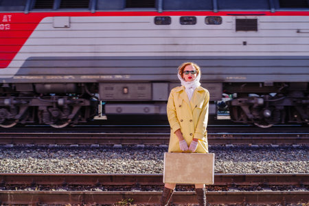 A young woman in a yellow raincoat and dark glasses, with a suitcase in her hands, stands on the...