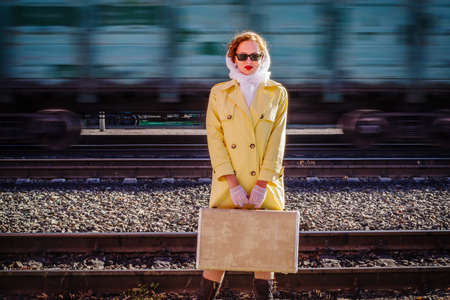 A young woman in a yellow raincoat and dark glasses, with a suitcase in her hands, stands on the platform of the railway station. Retro style outdoor shotの写真素材