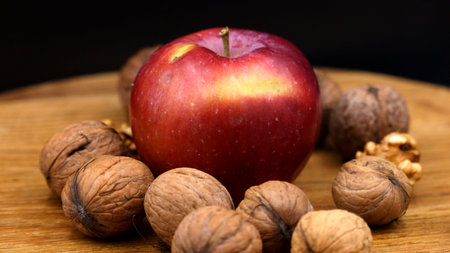 A vibrant red apple with natural blemishes is surrounded by whole walnuts on a warm wooden surface, captured with soft lighting. Perfect for healthy eating blogs, grocery ads, recipe books, and autumn-themed marketing campaigns.の写真素材