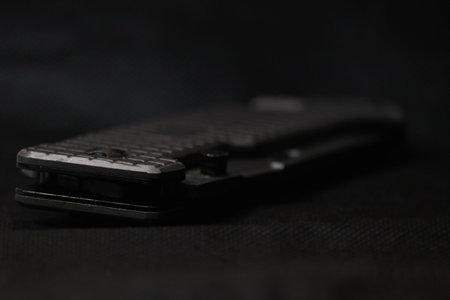 A modern black pocket knife with a textured handle is captured in a close-up, lying on a dark,...