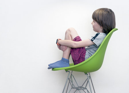 A pensive boy in casual wears is sitting on a green chair in front of a white wall.の写真素材