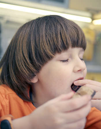 Caucasian boy with long light hair is eating chocolate sitting at the table in casual clothes....