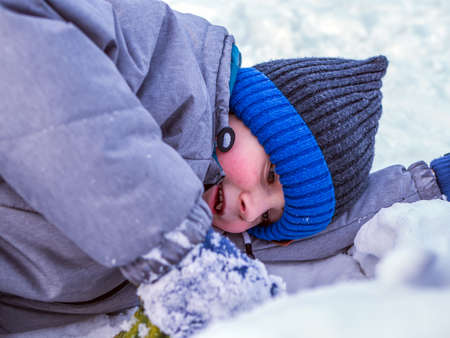 Smiling caucasian boy in green jacket and blue knit cap is lying on the snow.