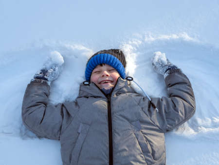 Smiling caucasian boy in green jacket and blue knit cap is lying on the snow.