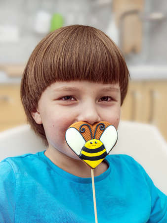 An eight-year-old caucasian boy in casual wear is holding a heart-shaped gingerbread in front of his...