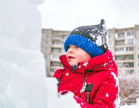 Caucasian boy in a red jacket and knitted hat plays in the snow in the city.
