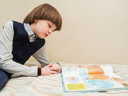 A boy in a school uniform reads a book at home while lying on the bed.