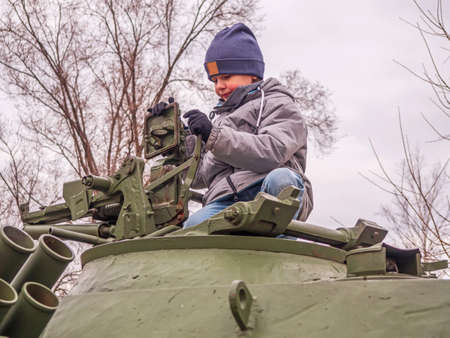 An eight-year-old Caucasian boy in a gray jacket and a blue knitted hat sits on a tank turret and is...