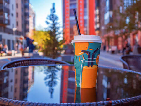 A beverage in a paper cup with a straw on a table of a street cafe. Close-up. Blurred background.