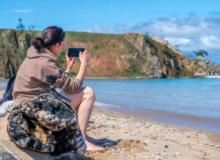 Video blogger girl is sitting on a wild shore and using a smartphone and an action camera.の写真素材
