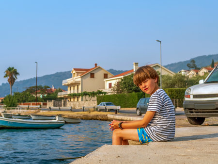 A boy in a striped t-shirt and swimming trunks is sitting near the seashore and looks at the camera.の写真素材