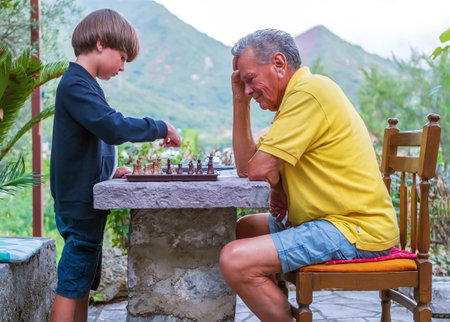 The boy is playing chess on the stone table with his grandfather in the backyard. Outdoors activity, outdoors leisure, board game.の写真素材