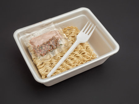 Dried block of instant noodle with seasoning sachets and plastic fork in a polystyrol lunch box package on the dark background. Content of a instant noodle box.の写真素材