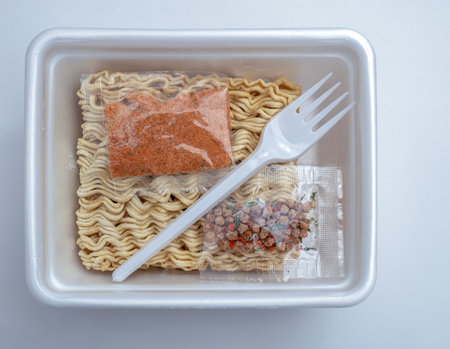 Dried block of instant noodle with seasoning sachets and plastic fork in a polystyrol lunch box package on the whie background. Content of a instant noodle box.の写真素材