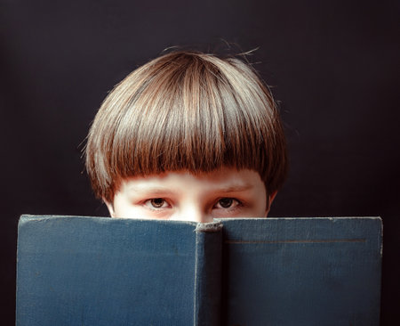 Caucasian boy looking at the camera covering the lower part of his face with a book. close-up.の写真素材