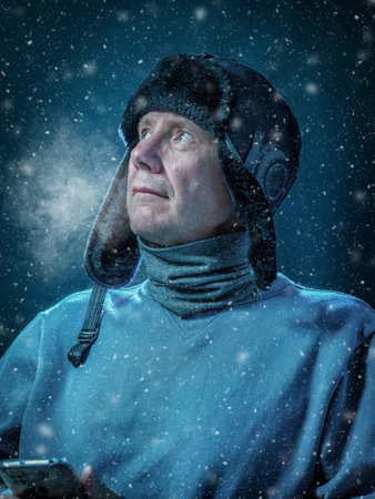 Caucasian man in warm winter clothes with a smartphone in his hands sitting in a dark and cold home. Vapor of breath. Concept of cold winter and high cost of heating.の写真素材