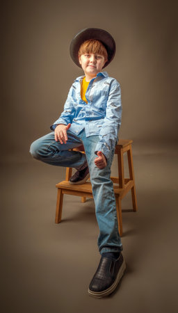 A 10-year-old boy in casual clothes and a fedora hat is sitting on a wooden chair and looking at camera.の写真素材