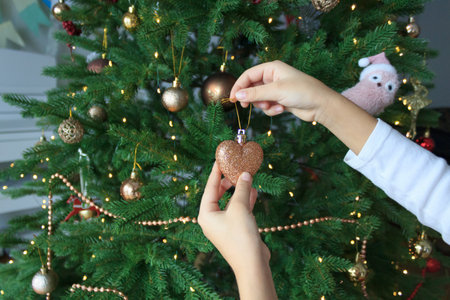 The child's hands hold a Christmas tree decoration in the form of a shiny heart against the background of a decorated Christmas tree.の写真素材