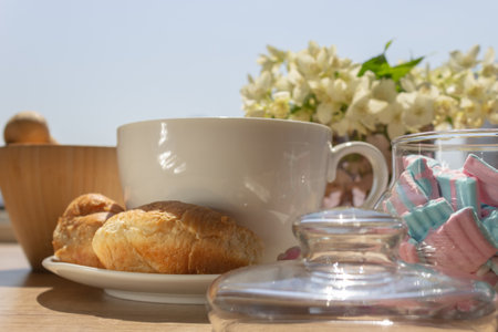Tea in a large white cup with fresh appetizing croissants, pink and blue delicate marshmallows in the sunlight against the background of flowers at an open windowの写真素材