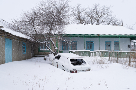 A snowy village yard during a snow storm with an abandoned car in the middle of it.の写真素材