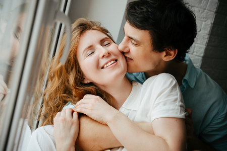 Happy girl and man kissing near window in home.の写真素材