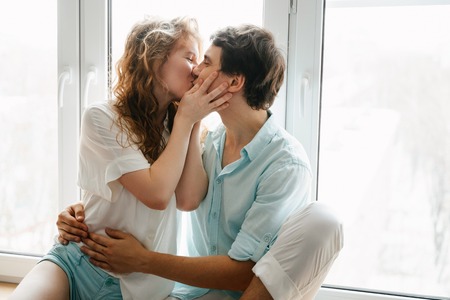 Happy girl and man kissing near window in home. White and blue clothes. Valentine day.の写真素材