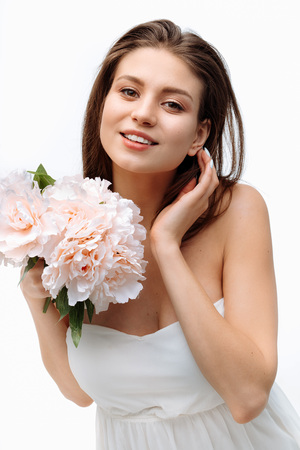 Portrait beautiful young girl smiling and posing with artificial flowers on white background in white dress.の写真素材