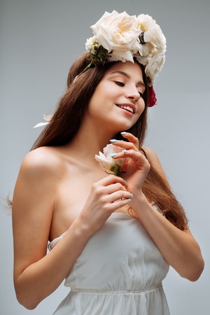 Beautiful young girl smiling and posing with flowers on gray background in white dress. Studio portrait.の写真素材
