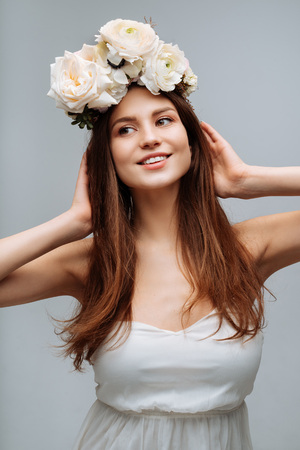 Beautiful young girl smiling and posing with flowers on gray background in white dress. Studio portrait.の写真素材