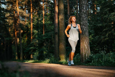 Young curly-haired woman runs along the forest path of the park and smiles. Happy sportswoman is engaged in fitness.の写真素材