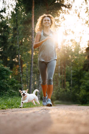 Young happy woman doing weight loss fitness in the park, running with her dog jack russell.の写真素材