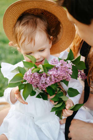 Happy smiling baby girl sniffing fresh spring flowers of lilac. The blonde daughter is sitting in the arms of her parents with flowers.の写真素材