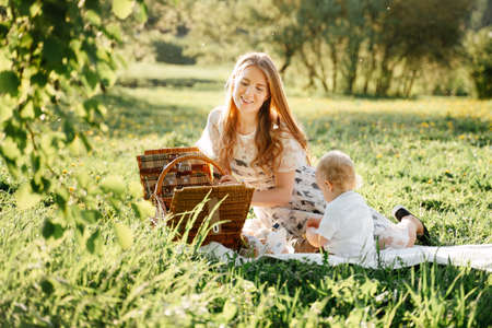 Happy mom with little daughter sits on a green lawn in the park on a picnic with baskets. Family day of parents with children.の写真素材