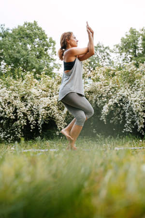 Full length side view of an athletic woman standing on one leg practicing yoga pose in a green park.の写真素材