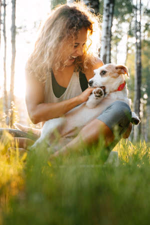 Happy smiling woman playing with her dog jack russell in the park while sitting on the grass.の写真素材