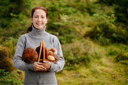 Young woman looking at the camera holds edible mushrooms collected in a basket against the background of a green forest in summer.の写真素材