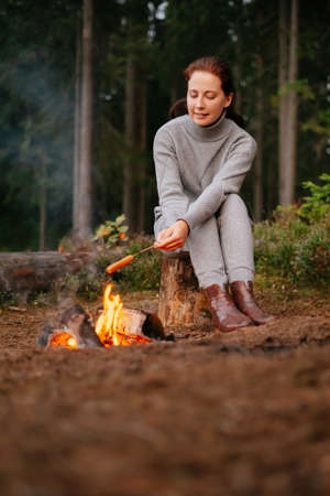 A tourist woman at a campsite is cooking meat food on an open fire in the forest. Concept of a hike and ecotourism.の写真素材