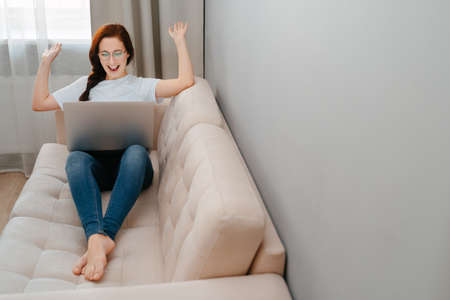 Joyful young woman celebrates winning an online lottery while sitting at home on a white sofaの写真素材