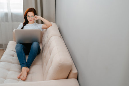 Young woman with glasses is sitting on a bright sofa watching movie and working on laptop.の写真素材