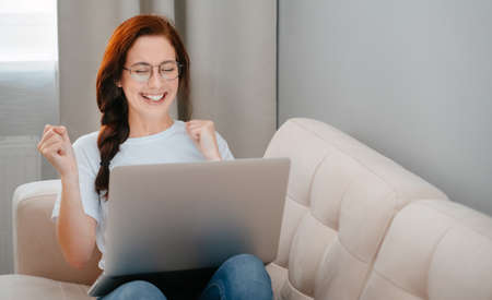 Young woman enjoys success looking at the laptop screen while sitting on a cozy sofa in the living room of the house.の写真素材